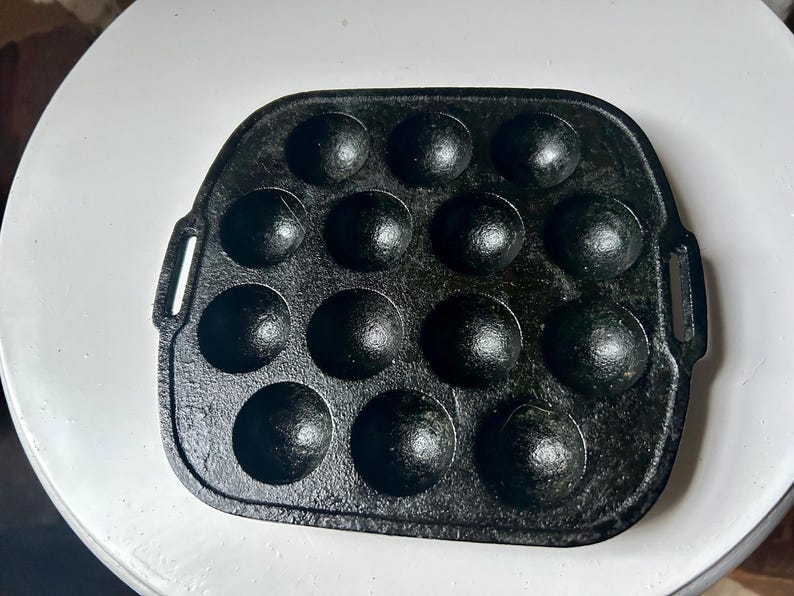 May include: A black cast iron takoyaki pan with 14 spherical moulds. The pan has a rectangular shape with rounded corners and small handles on each side. The pan is on a white surface.