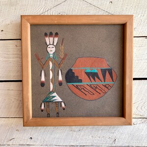 Puede incluir: Una pintura de arena enmarcada que representa una figura nativa americana con un vestido tradicional, un tocado y una olla con acentos turquesa y negros.