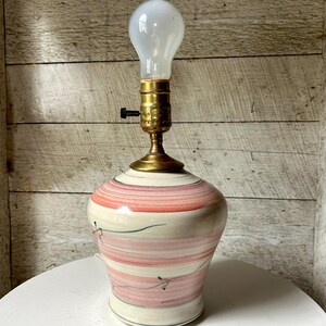 May include: A ceramic table lamp with a bulb. The lamp base is a cream color with horizontal stripes of pink and red. The lamp has a brass neck and a white bulb. The lamp is on a white surface.