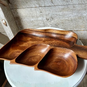 Könnte beinhalten: Ein Holzserviertablett mit einem blattartigen Design. Das Tablett ist in drei Abschnitte unterteilt und hat einen Griff. Das Holz hat eine satte, dunkelbraune Farbe und sichtbare Maserung. Das Tablett wird wahrscheinlich zum Servieren von Snacks oder Vorspeisen verwendet.