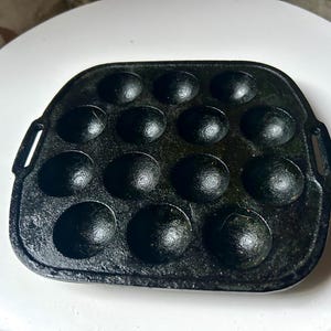 May include: Black cast iron takoyaki pan with 14 hemispherical moulds. The pan has a rectangular shape with rounded corners and two small handles on the sides. The pan is designed for cooking Japanese takoyaki.