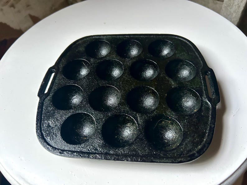 May include: Black cast iron takoyaki pan with 14 hemispherical moulds. The pan has a rectangular shape with rounded corners and two small handles on the sides. The pan is designed for cooking Japanese takoyaki.