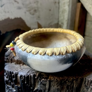 May include: A handcrafted, silver-colored bowl with a decorative, light brown rim. The rim has a scalloped edge. A small, colorful fishing lure is attached to the side of the bowl. The bowl is sitting on a weathered, dark brown wooden surface.