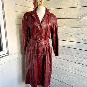 Puede incluir: Un abrigo largo de cuero burdeos con cinturón en la cintura y cierre de botones. El abrigo tiene mangas largas, cuello y dos bolsillos delanteros. El abrigo se exhibe en un maniquí de madera.