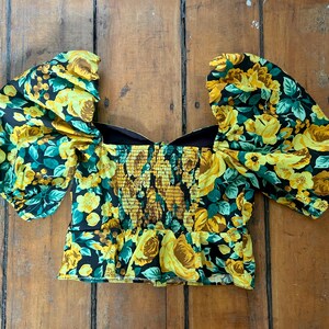 May include: A floral print top with a square neckline and puffed sleeves. The top features a yellow rose pattern on a black background, with a fitted, smocked bodice. The garment is made of a lightweight fabric.