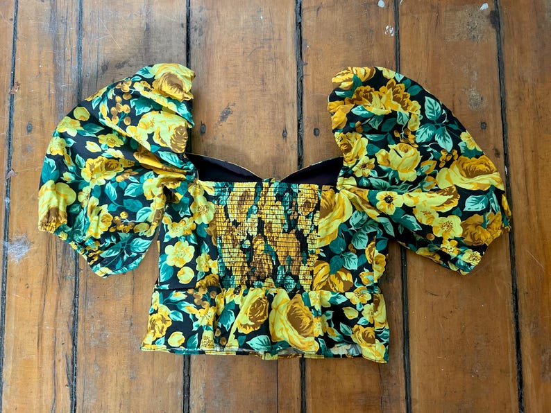 May include: A floral print top with a square neckline and puffed sleeves. The top features a yellow rose pattern on a black background, with a fitted, smocked bodice. The garment is made of a lightweight fabric.