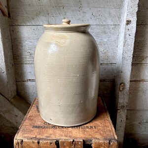 May include: A tall, cylindrical stoneware crock with a smooth, light beige glazed finish and a matching fitted lid. The vessel features a slightly tapered neck and a small, rounded knob handle on the center of the lid, presenting a simple, functional design.