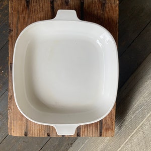 May include: White ceramic baking dish with two handles. The dish is empty and sitting on a wooden surface.
