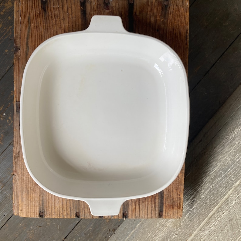 May include: White ceramic baking dish with two handles. The dish is empty and sitting on a wooden surface.