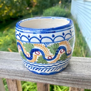 May include: A ceramic planter with a hand-painted design. The planter features a white interior and a blue, green, and yellow exterior pattern. The design includes wavy lines, leaf shapes, and dots. The planter is cylindrical and sits on a wooden surface.