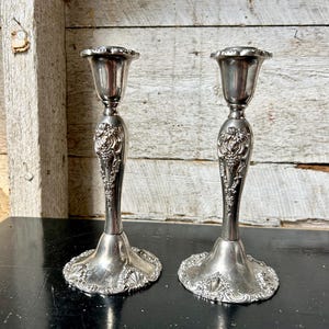 Könnte beinhalten: Zwei silberne Kerzenständer mit aufwendigen floralen und Weinreben-Details. Die Kerzenständer haben eine gewellte Oberseite und einen breiten, dekorativen Sockel. Der Hintergrund ist eine rustikale Holzwand.