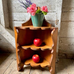 May include: A three-tiered wooden corner shelf with scalloped edges. The top shelf holds a light green ceramic pot containing pink artificial flowers and purple sprigs. A shiny red apple sits on each of the two lower shelves of the unit.