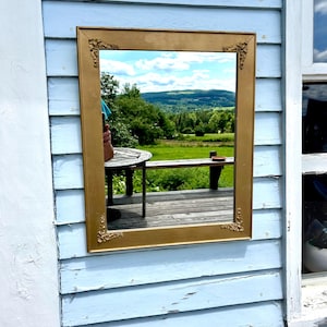 May include: A rectangular gold-framed mirror reflecting a scenic outdoor view of a green field, trees, and a blue sky with clouds. The mirror is mounted on a light blue wooden wall.