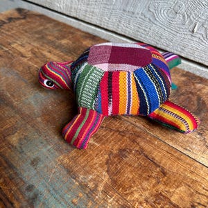 May include: A colorful patchwork fabric turtle toy. The turtle is made of different colored stripes and plaid fabric. The turtle is lying on a wooden surface.