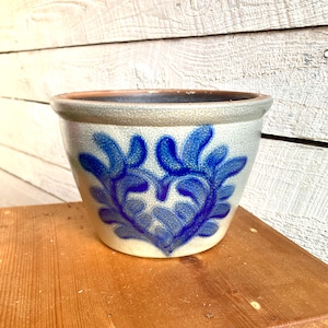 Puede incluir: Una maceta de cerámica blanca con un diseño azul en forma de corazón. La maceta tiene una superficie rugosa y texturizada y una abertura ancha y redonda.