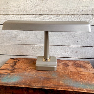Puede incluir: Una lámpara de escritorio vintage de color plateado con una pantalla larga y rectangular y una base redonda. La lámpara está sobre una superficie de madera.