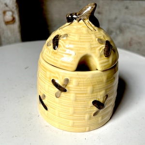 May include: A ceramic honey pot with a lid, shaped like a beehive. The pot is light yellow with a woven texture and features several painted bees. The lid has a small opening and a brown bee-shaped handle.