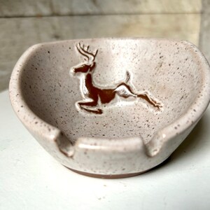 Könnte beinhalten: Ein weißer Aschenbecher aus Keramik mit einem braunen Hirsch-Design. Der Aschenbecher hat eine gesprenkelte Glasur und eine kleine Vertiefung für Zigaretten.