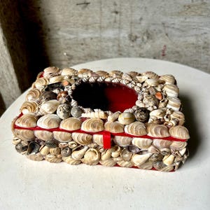 Puede incluir: Una caja cuadrada roja cubierta de pequeñas conchas. La caja tiene un pequeño espejo redondo rojo en el interior.