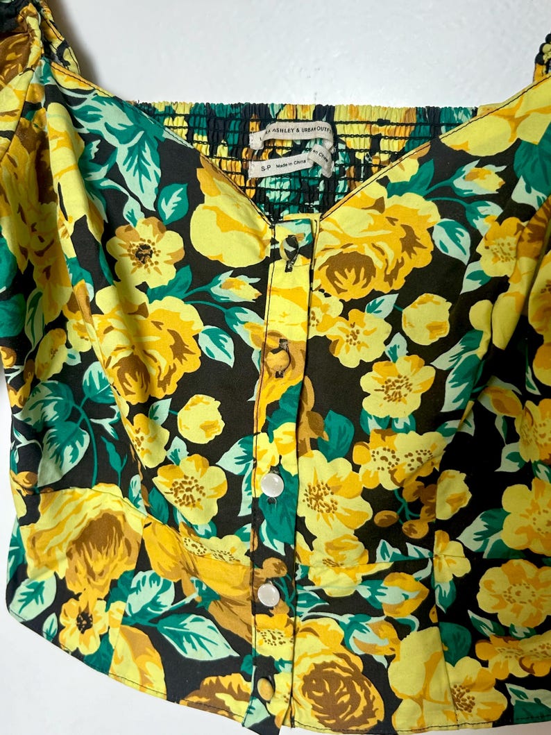 May include: A floral print top with yellow roses, green leaves, and a black background. The top has a square neckline, button-up front, and a smocked back. The tag reads "ASHLEY & URBAN OUTFITTERS".