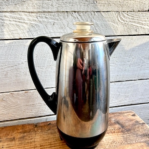 May include: A vintage stainless steel coffee pot with a black handle and base. The pot has a silver body with a black base and handle. The lid is a light beige color. The coffee pot is sitting on a wooden surface.