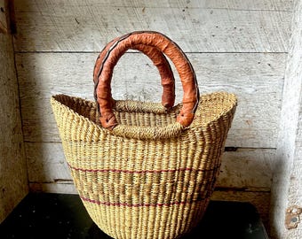 Puede incluir: Una cesta tejida con forma redondeada y dos asas de cuero marrón. La cesta está hecha de fibras de color natural con finas rayas rojas. Las asas son de un tono marrón más oscuro.
