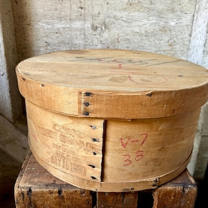 May include: A round, light brown wooden box with a lid. The box has a cylindrical shape and is made of thin wooden planks held together with small nails. The side of the box has faded text and the numbers "V-7" and "38" in red.
