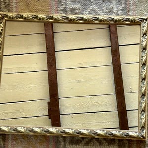 May include: A rectangular mirror with a decorative gold-toned frame featuring a repeating leaf pattern. The mirror reflects a light-colored wooden wall made of horizontal planks and two vertical dark brown wooden support beams.