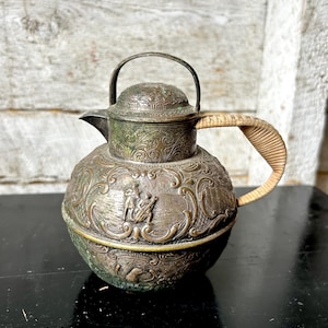 May include: Antique silver-toned metal teapot with intricate floral and figural relief details. Features a woven handle and a hinged lid with a small handle. The teapot has a spout and a rounded body.