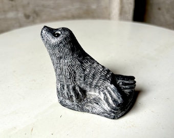 May include: A small, gray stone sculpture of a seal looking up. The seal is carved in a realistic style with detailed fur and a textured surface.