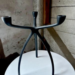 May include: A black metal candelabra with three candle holders. The candelabra has an abstract design with three legs. The candle holders extend horizontally from a central stem. The piece is displayed on a white surface.