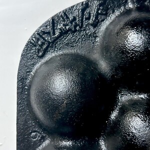 May include: Close-up of a black cast iron mould with a textured surface. The mould features several rounded cavities and a raised inscription along the edge. The design suggests a baking or cooking tool.