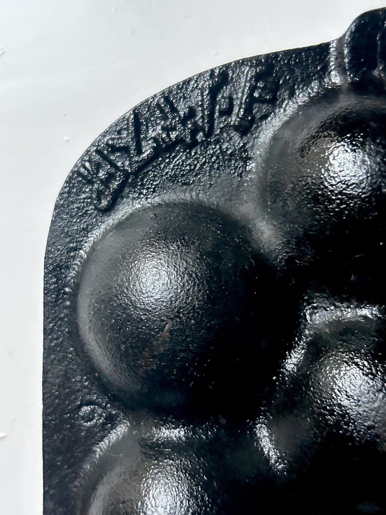 May include: Close-up of a black cast iron mould with a textured surface. The mould features several rounded cavities and a raised inscription along the edge. The design suggests a baking or cooking tool.