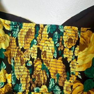 May include: Close-up of a floral print dress featuring yellow roses, green leaves, and brown accents on a black background. The bodice has a smocked design. The dress has a sweetheart neckline and wide straps.