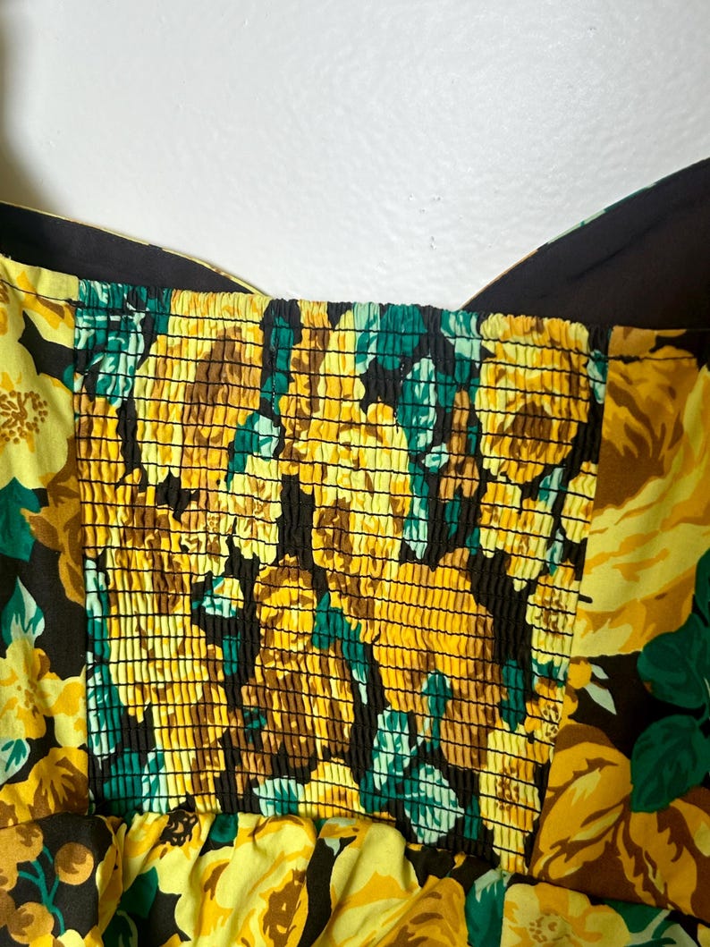 May include: Close-up of a floral print dress featuring yellow roses, green leaves, and brown accents on a black background. The bodice has a smocked design. The dress has a sweetheart neckline and wide straps.
