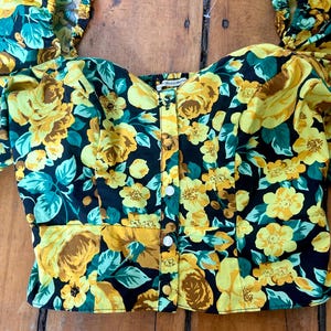 May include: A floral print top with a black background, yellow roses, and teal leaves. The top has a button-down front and short, puffed sleeves. The fabric is a lightweight material.