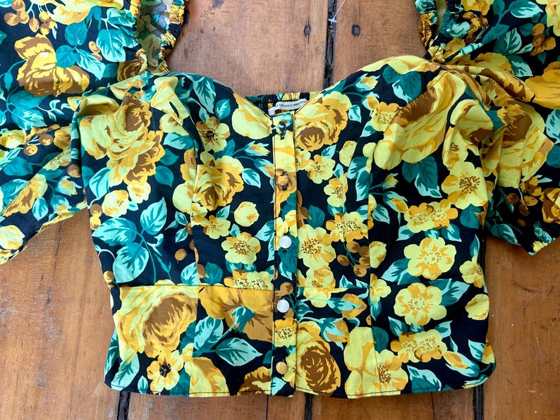 May include: A floral print top with a black background, yellow roses, and teal leaves. The top has a button-down front and short, puffed sleeves. The fabric is a lightweight material.