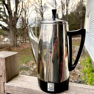 Może przedstawiać: Elektryczny ekspres do kawy ze stali nierdzewnej z czarną podstawą i uchwytem. Ekspres do kawy ma błyszczącą, odblaskową powierzchnię i czarną pokrywę. Nazwa marki "Presto" jest widoczna na podstawie. Ekspres do kawy stoi na drewnianej powierzchni.