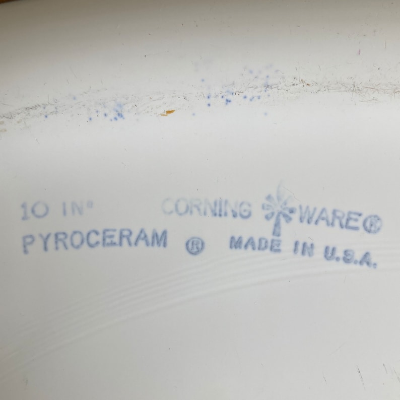 May include: A white 10-inch Corning Ware Pyrex baking dish with the text "Corning Ware" and a stylized palm tree logo. The dish also has the text "Pyroceram" and "Made in U.S.A."