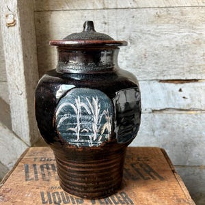 May include: A dark brown ceramic jar with a lid. The jar has a cylindrical base and a wider body with a circular design featuring white and green plants. The lid is a darker shade of brown. The jar is sitting on a wooden crate.