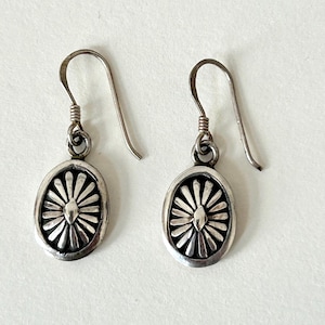 May include: A pair of silver-toned earrings. Each earring features an oval-shaped pendant with a sunburst design. The earrings have a hook closure. The earrings are displayed on a white background.