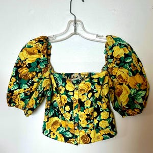 May include: A floral print top with a square neckline and short, puffed sleeves. The fabric features yellow roses and green leaves on a black background. The top is buttoned down the front and hangs on a clear hanger.