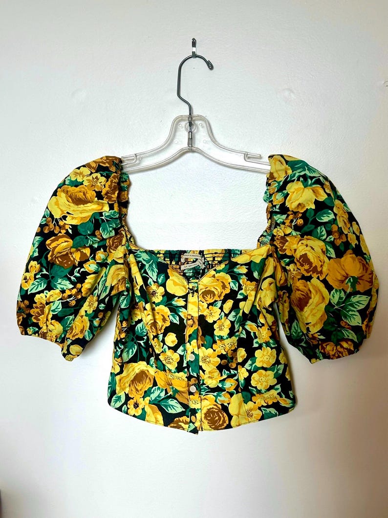 May include: A floral print top with a square neckline and short, puffed sleeves. The fabric features yellow roses and green leaves on a black background. The top is buttoned down the front and hangs on a clear hanger.