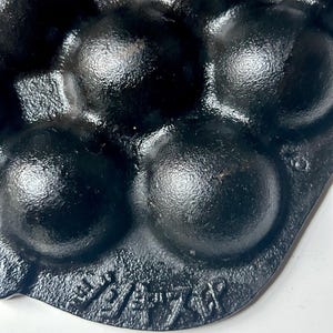 May include: Close-up of a black cast iron muffin pan with multiple rounded moulds. The pan has a textured surface and is marked with embossed text. The pan is designed for baking muffins or similar treats.
