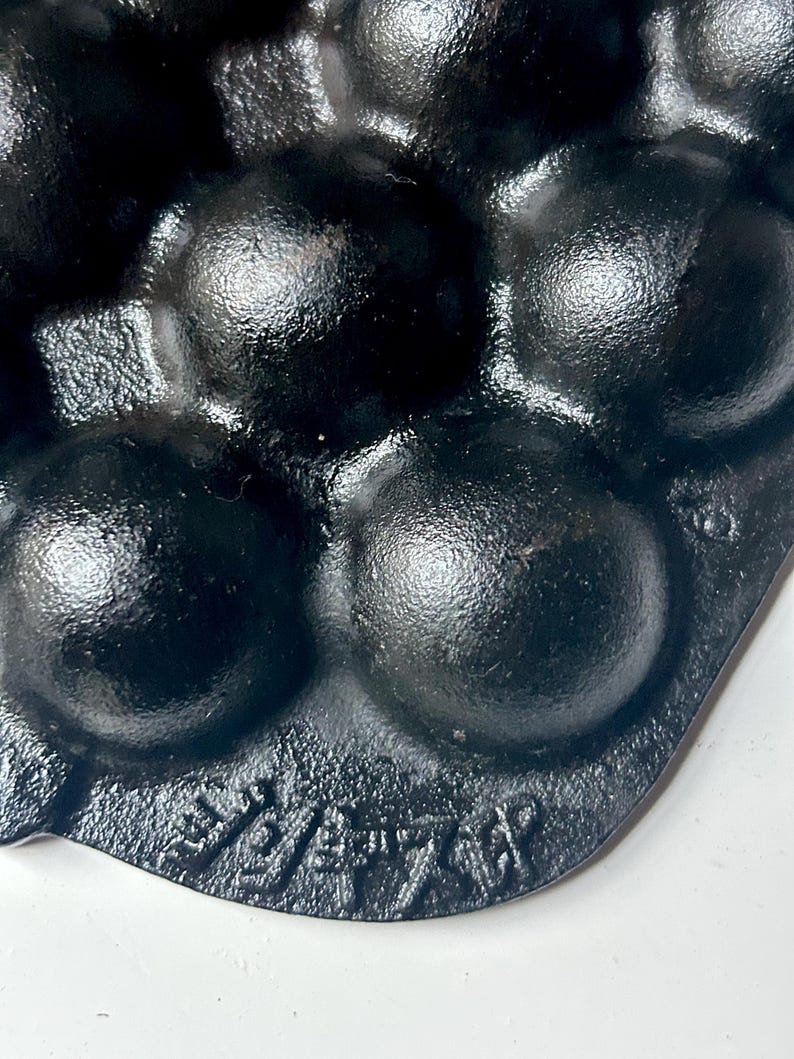 May include: Close-up of a black cast iron muffin pan with multiple rounded moulds. The pan has a textured surface and is marked with embossed text. The pan is designed for baking muffins or similar treats.