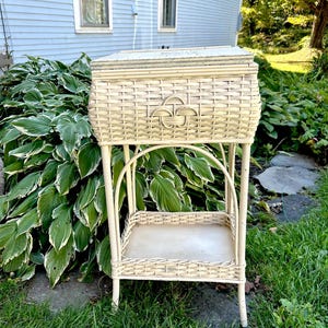 May include: An antique cream-coloured wicker sewing basket on a stand. The basket has a decorative front design and a hinged lid. The stand features a lower shelf and four legs, all in a matching cream colour. The basket is set outdoors in a garden.