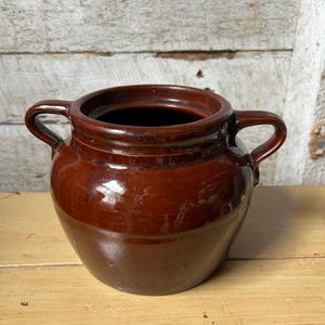 May include: A brown ceramic jar with two handles. The jar has a glossy finish and a wide opening. The color is a rich, dark brown, with a slightly lighter shade on the lower portion. The jar is likely used for storage or decoration.