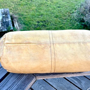 May include: A large, rectangular, tan leather bag with a rounded shape. The bag has a stitched seam down the center and is resting on a wooden surface. The leather has a worn appearance.