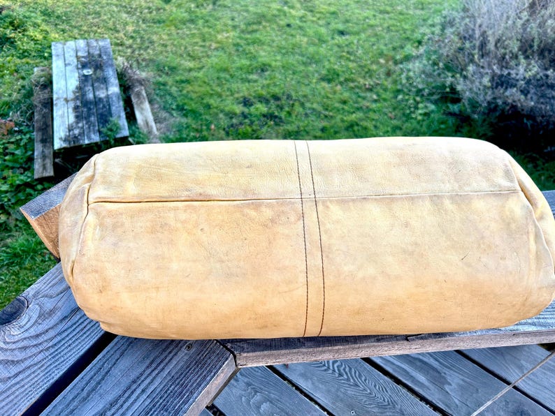 May include: A large, rectangular, tan leather bag with a rounded shape. The bag has a stitched seam down the center and is resting on a wooden surface. The leather has a worn appearance.