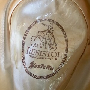 May include: A brown and white Resistol Western hat with a cowboy riding a horse logo. The hat is labeled "Self-Conforming".
